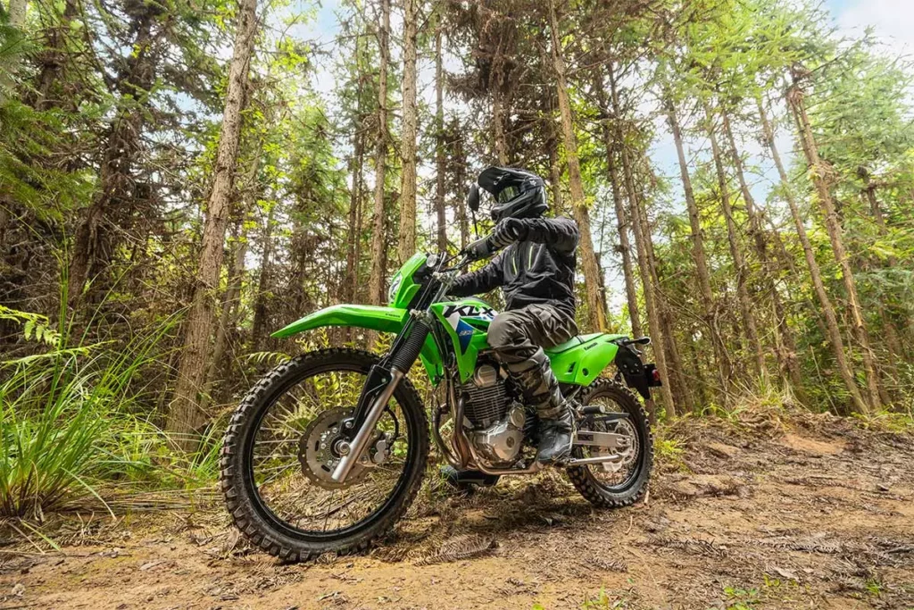 Piloto conduz Kawasaki KLX230 S verde por trilha entre árvores.
